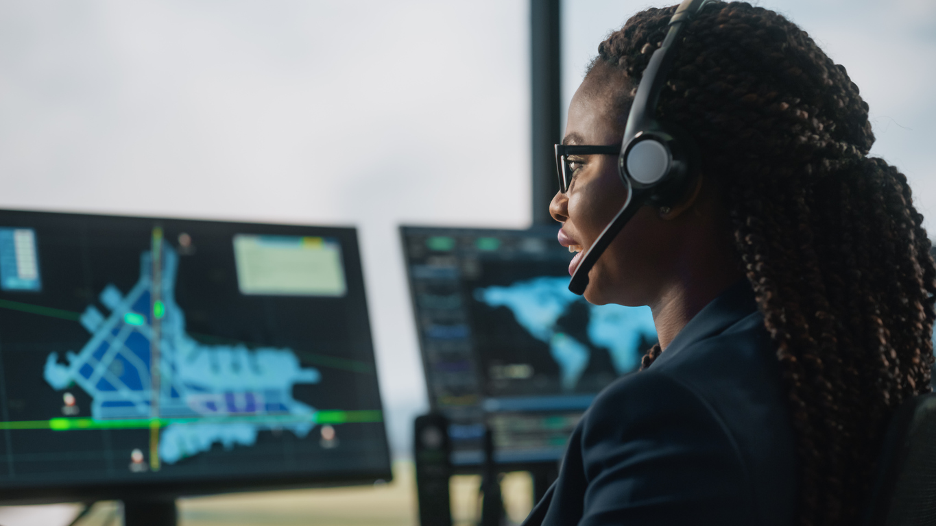 Female Air Traffic Controller with Headset Talk on a Call in Airport Tower. Office Room is Full of Desktop Computer Displays with Navigation Screens, Airplane Departure and Arrival Data for the Team.