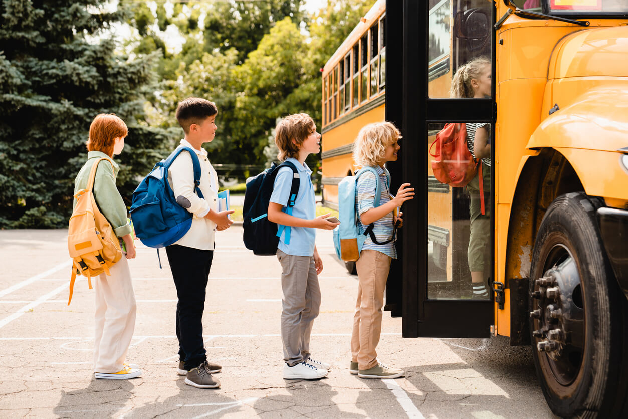 Multiethnic mixed-race pupils classmates schoolchildren students standing in line waiting for boarding school bus before starting new educational semester year after summer holidays