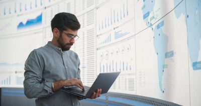 Indian Male Product Distribution Expert Holding Laptop Computer, Analyzing World Map On Big Digital Screen In Monitoring Office. Successful Man Looking For Efficient Routes For International Delivery.