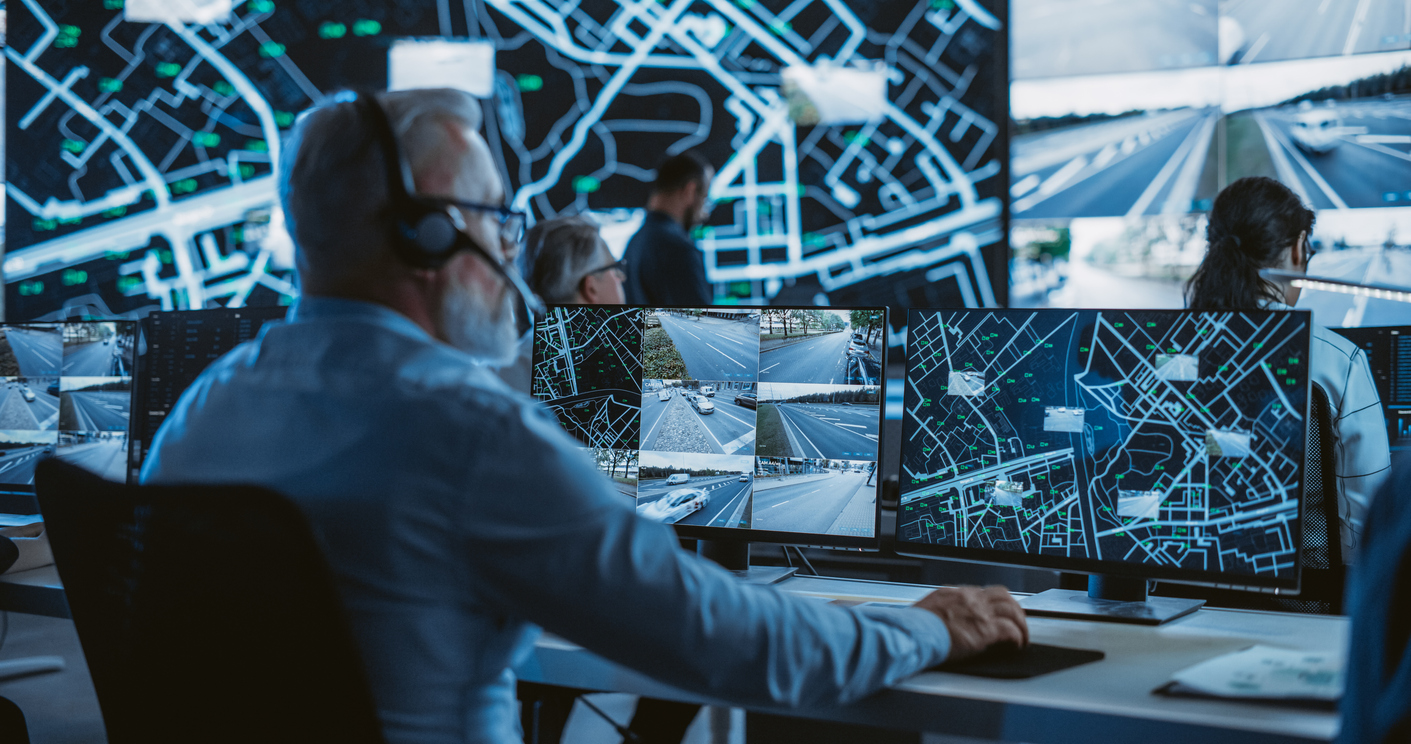 Technical Customer Support Specialist is Talking on a Headset while Working on a Computer in a Call Center Control Room Filled Display Screens with Monitoring Software. Over the Shoulder Footage